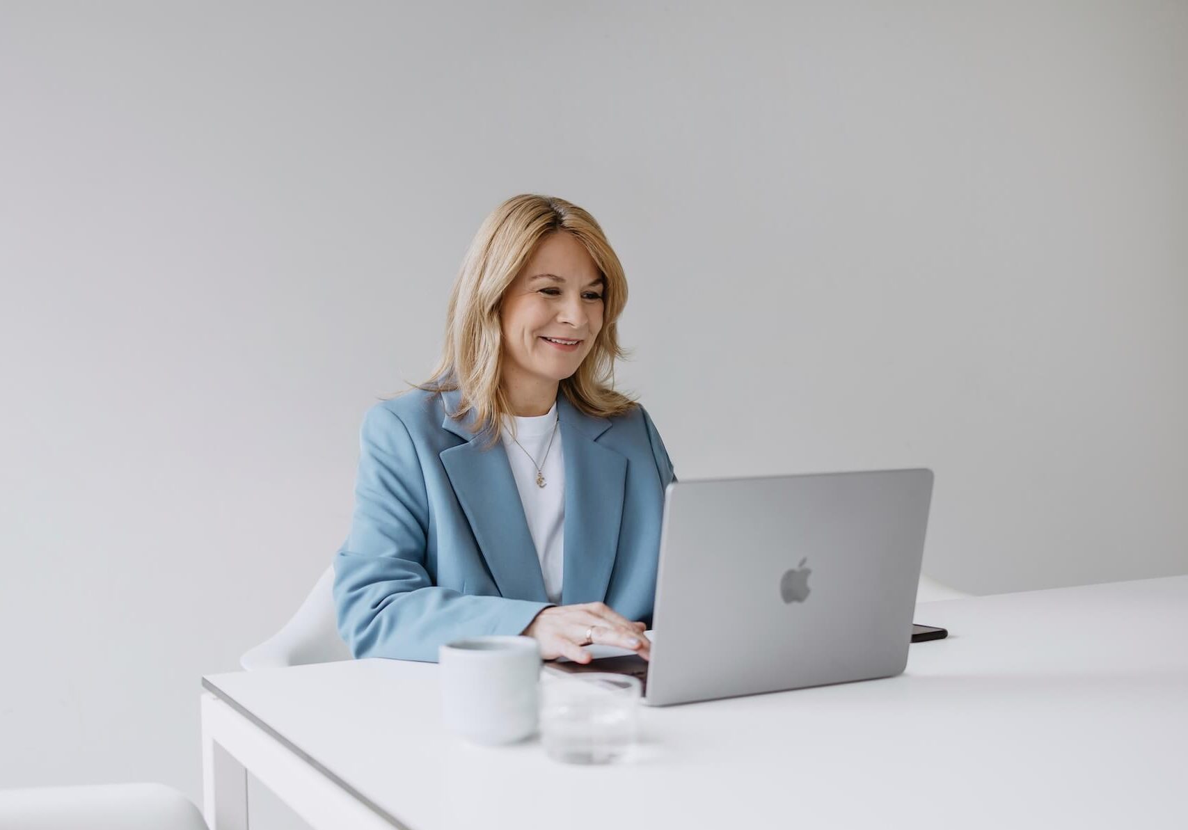 Blonde Frau sitzt in einem weißen Raum vor einem Macbook